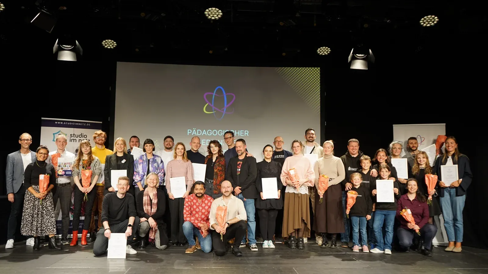 Gruppenfoto aller fünfzehn ausgezeichneten Teams und Studios des Pädagogischen Medienpreises 2025 auf der Bühne im Gasteig HP8 München