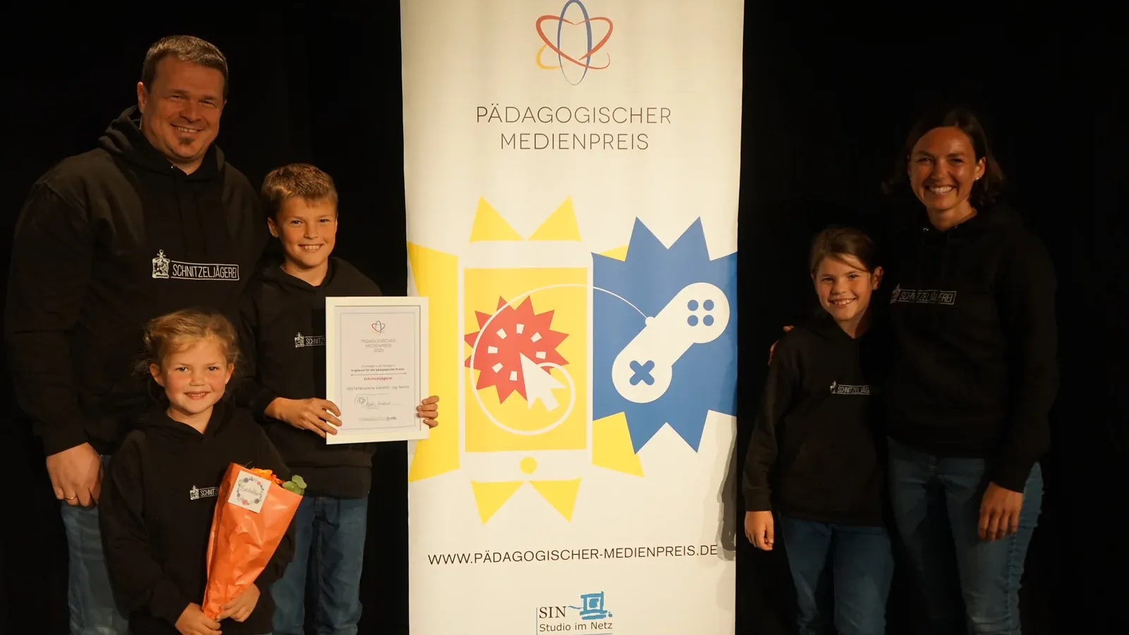 Schnitzeljägerei-Team im einheitlichen Hoodie steht mit Urkunde und Blumenstrauß vor dem Roll-up des Pädagogischen Medienpreises