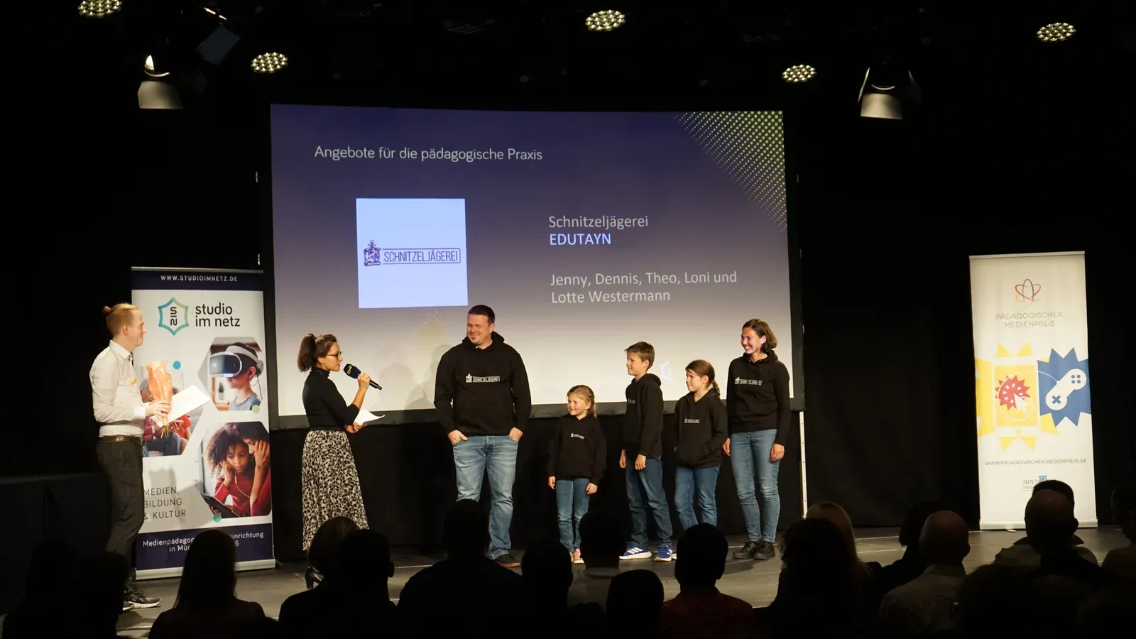 Schnitzeljägerei-Team steht auf der Bühne im Gasteig HP8 — Moderation hält Mikrofon, im Hintergrund die Slide mit dem Schnitzeljägerei-Logo und der Kategorie ‚Angebote für die pädagogische Praxis‘