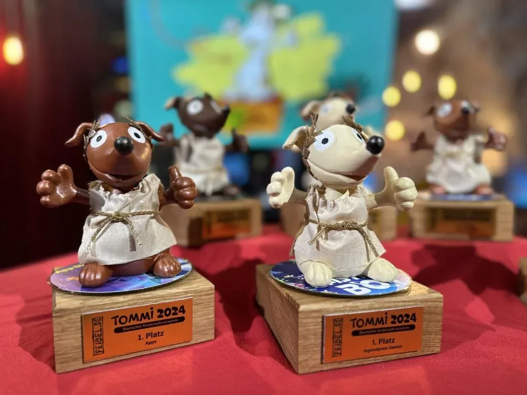 Die TOMMI-2024-Trophäen auf rotem Tuch — Maskottchen-Figuren mit Lorbeer-Kranz auf Holzsockeln mit orangefarbenen Schildern